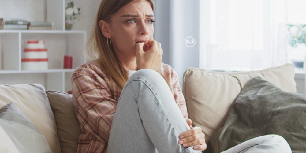 Mladá žena sedící na světlém gauči v obyvacím pokoji s rukou dramaticky opírající se o ústa a bradu druhou rukou se držící skrčenou nohu v presu signalizující, že žena prožívá stresi, napětí či depresi | Pro článek: Jak na depresi? | Pro web: radyzezivota.cz | Zdroj: shutterstock.com
