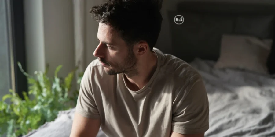 Mladý muž s natočenou hlavou do strany z profilu se zamyšlenou náladou spíše nešťastným pohledem do strany sedící v ložnici na posteli po levé straně okno za zády muže postel s polštáři | Pro článek: Jak na depresi? | Pro web: radyzezivota.cz | Zdroj: shutterstock.com