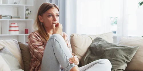 Mladá žena ve stresu sedící na gauči v obyváku s rukou na ústech a druhou rukou držící si skřížené nohy upřeně se dívající v tlaku dopředu | Pro článek: Jak na prázdnotu | Pro web: radyzezivota.cz | Autor úpravy fotografie: Jiří Samuel | Zdroj: shutterstock.com