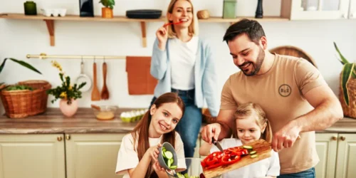 Rodina v kuchyni společně připravující salát z čerstvé zeleniny z paprik a okurek na dřevěném prkénku v rukou nůž otce stahující nařezané kusy zeleniny do misky ležící na stole u kterého sedí 2 dcery jedna starší a jedna mladší a v pozadí stojící maminka dohlížející na průběh ochutnávající kus papriky v pozadí bílá stěna s vybavením kuchyně barevnější a trošku tmavší vzhled fotografie | Pro článek: Jídlo a návyky zásadním způsobem ovlivňují | Pro web: radyzezivota.cz | Zdroj: shutterstock.com