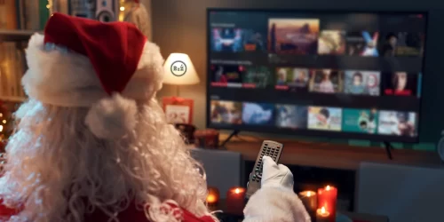 Santa Clause oblečený ve svém sedící v obyváku dívající se na televizi držící v ruce dálkový ovladač v pozadí příjemné prostředí obyváku a teplo domova se svítící lampou po levé straně po pravé straně TV s pořady | Pro článek: Nejlepší vánoční filmy - seznam vánočních pohádek | Pro web: radyzezivota.cz (Rady ze života) | Zdroj: shutterstock.com