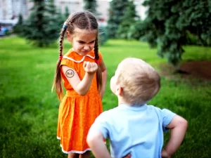 Fotografie mladé dívky držící pěstičku v horní části těla naznačující boj vůči svému mladšímu sourozenci chlapci v pozadí tráva a stromy s domy | Zdroj: shutterstock, web: radyzezivota | Článek: Výchova sourozenců